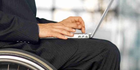 Image of a man wearing a suit, sitting in a wheelchair, and using a laptop computer.