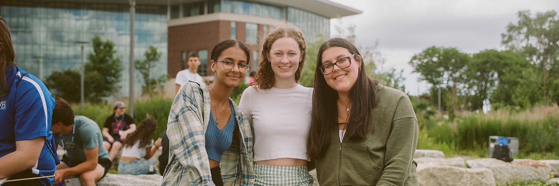 Three students smile at camera on campus lawn in front of university hall.