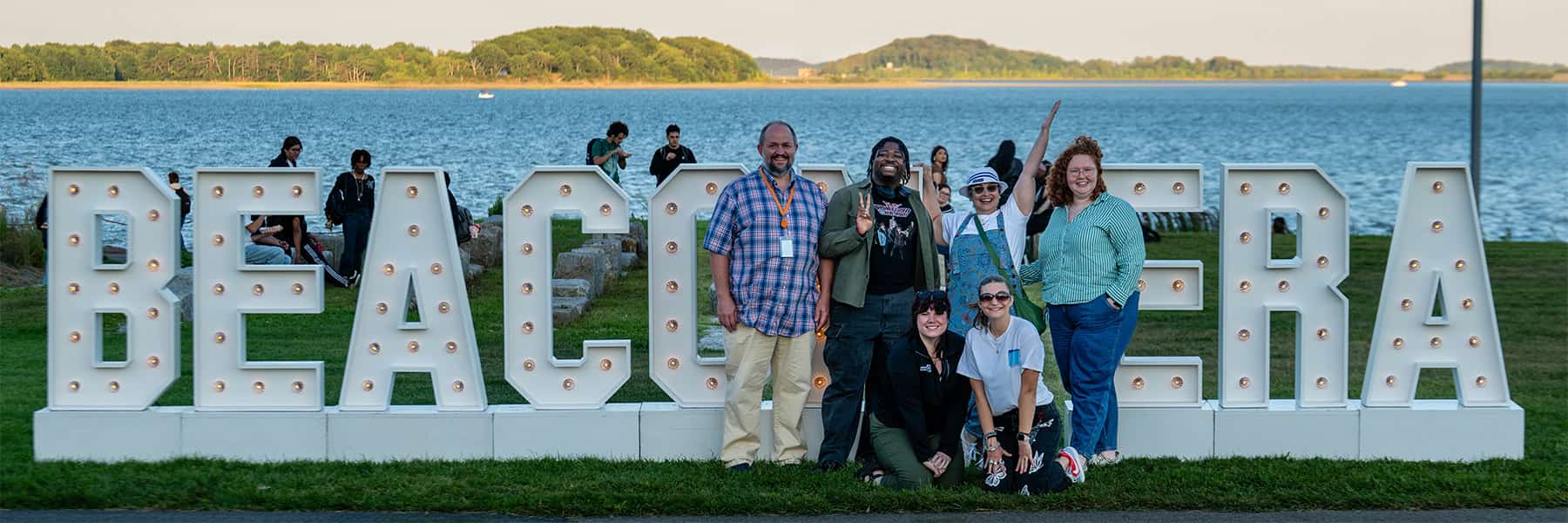 UMass Boston community posed in front of Beacon Era sign on the lawn with water in the background.