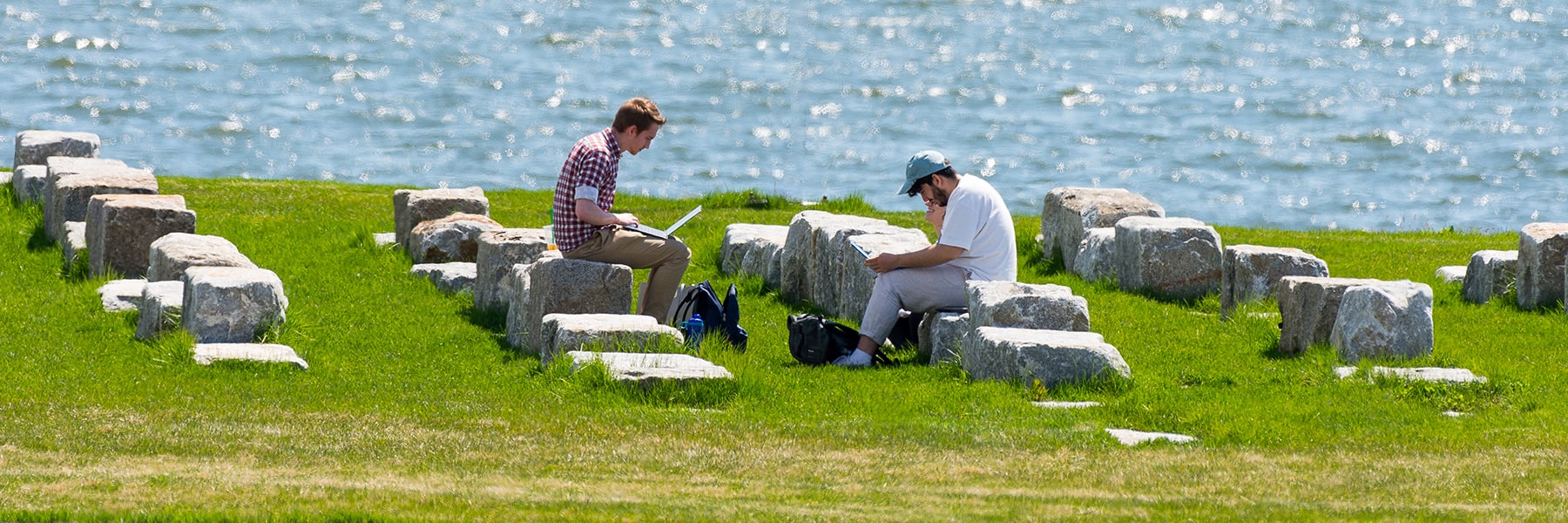 Students sit on stone benches by water on campus center lawn and work on laptops.