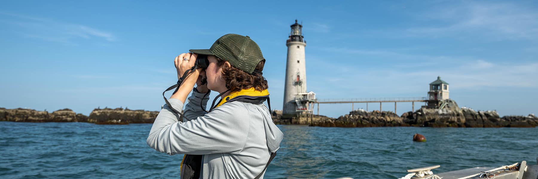 Student on a boat seal watching with binoculars with a lighthouse in the background.