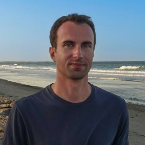 man with short brown hair and blue t-shirt standing in front of the beach