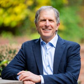 Man in blue suit with light gray hair, in the background there are shrubs and trees