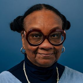 woman with dark hair in two buns, red round glasses and blue turtleneck in front of blue background