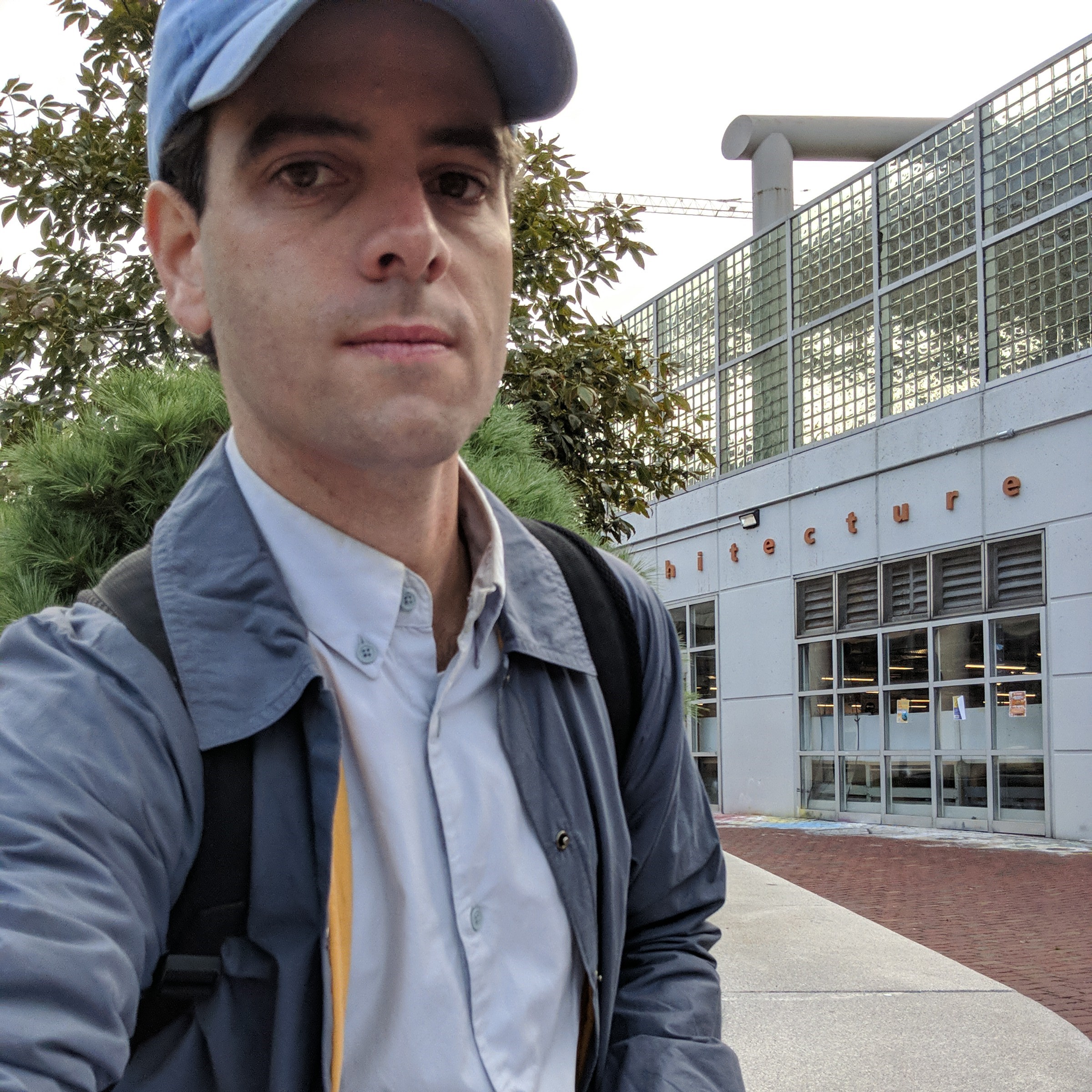 Man with blue baseball cap, grey jacket, in front of greenery and buildings