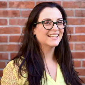 Woman with long brown hair, glasses, yellow shirt in front of brick wall