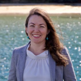 Woman with long light brown hair, white shirt, gray blazer, standing in front of water on a sunny day
