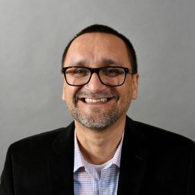 man with cropped dark hair, black glasses, black blazer and light shirt against grey background