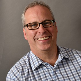 Man with short grey hair, rectangular glasses, and checkered button-up shirt against grey background